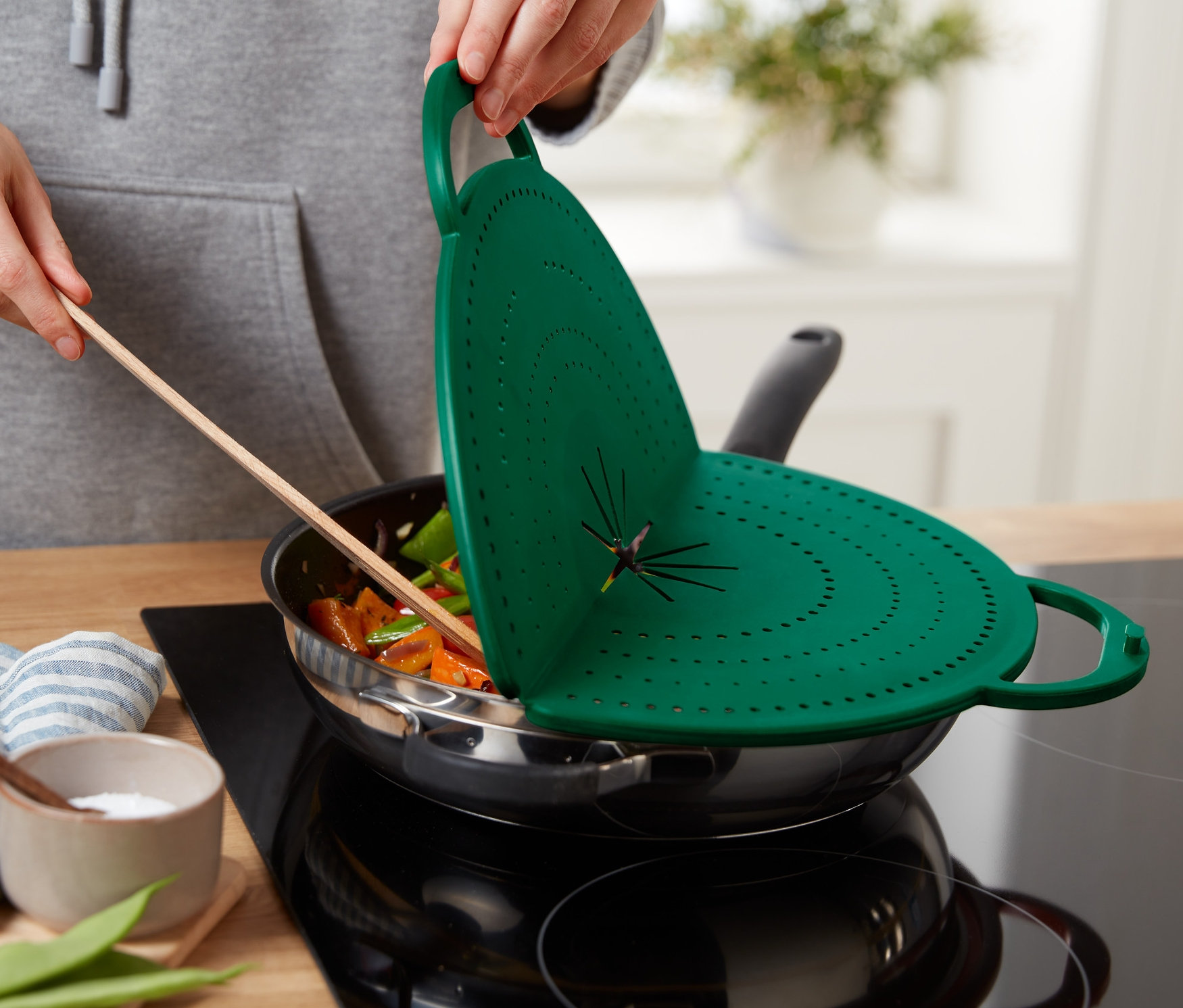 Bir kişi, içinde yemek olan bir tavanın üzerinde yeşil renkli 4'ü 1 Arada Yağ Sıçratmaz tutuyor.