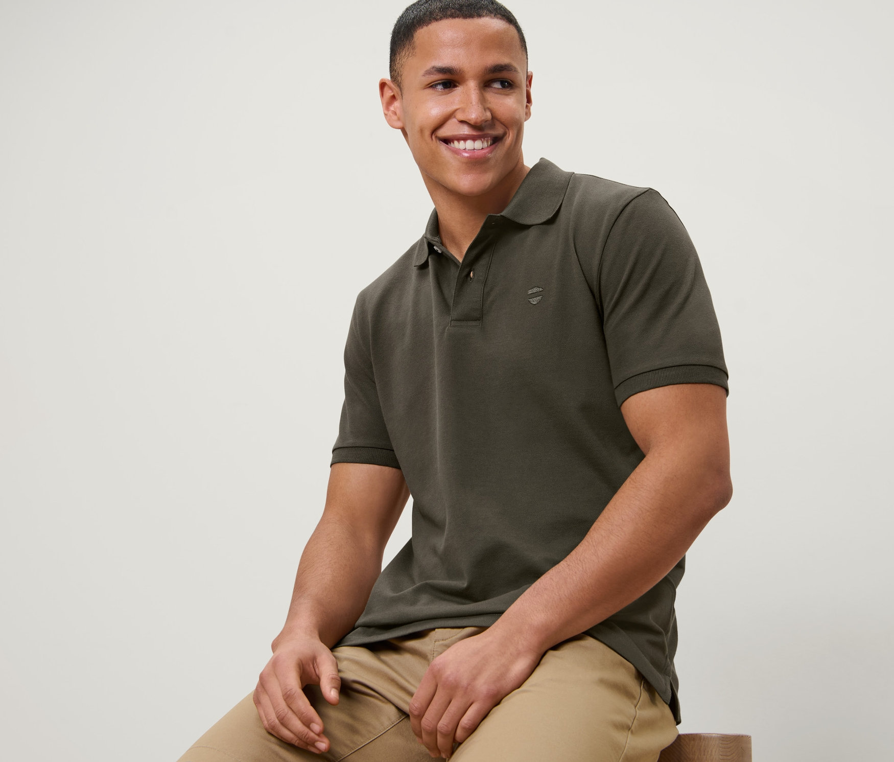 Bir adam taburede oturuyor ve zeytin rengi pike polo yaka tişört ve bej şort giyiyor.