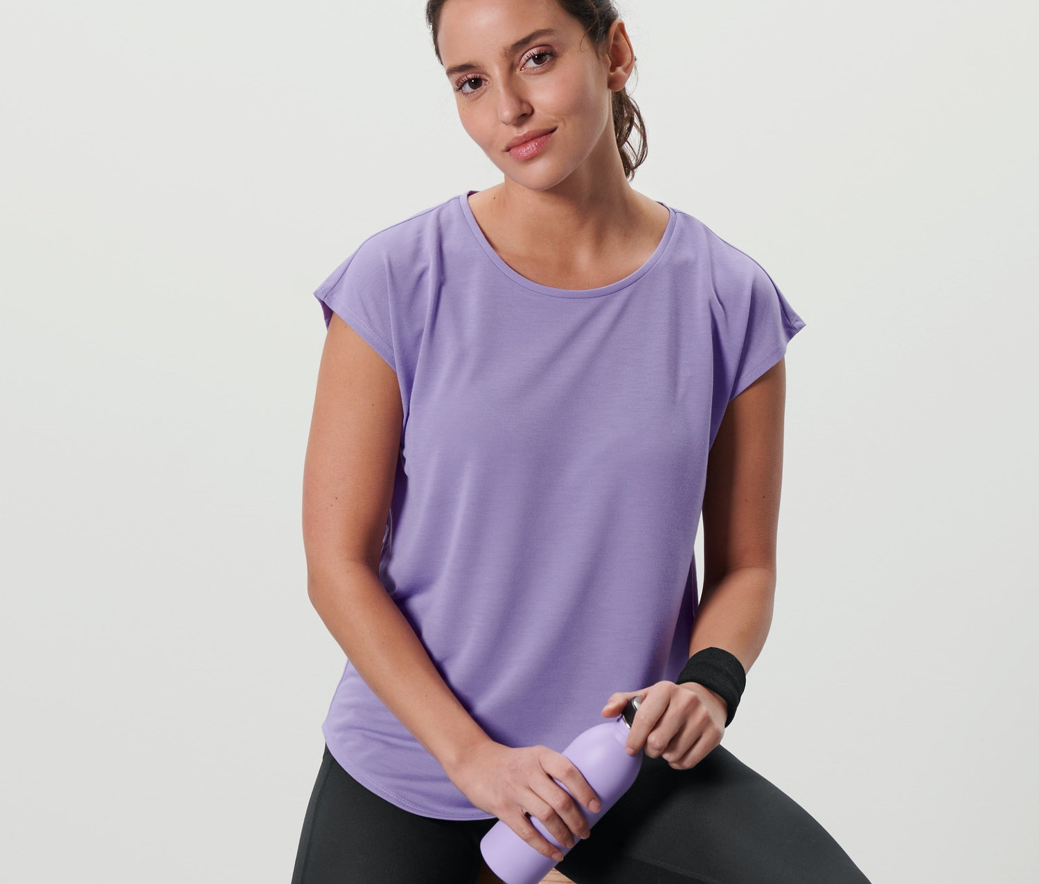 Kadın taburede oturuyor, lila rengi spor tişörtü giyiyor ve mor bir şişe tutuyor.