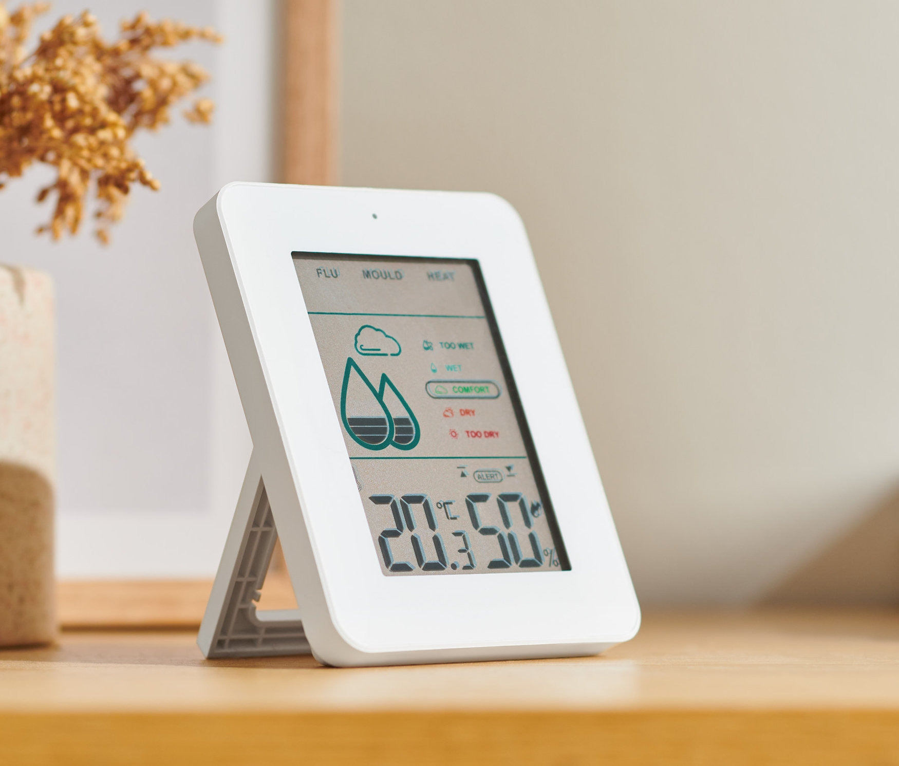 Küf alarmlı dijital nem ölçer, çiçek vazosunun yanında ahşap bir masa üzerinde duruyor.