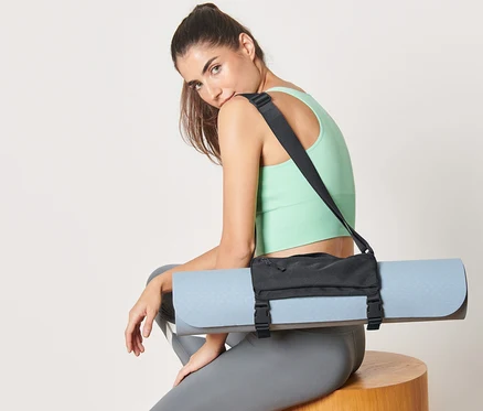 Kadın oturuyor, omzunda mavi bir yoga matı tutan bir mat taşıma kemeri var.