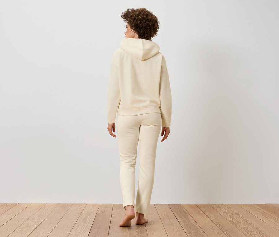 Kıvırcık saçlı bir kadın, ahşap zeminde bej kapüşonlu ve eşofman altı giyerek arkası dönük duruyor.