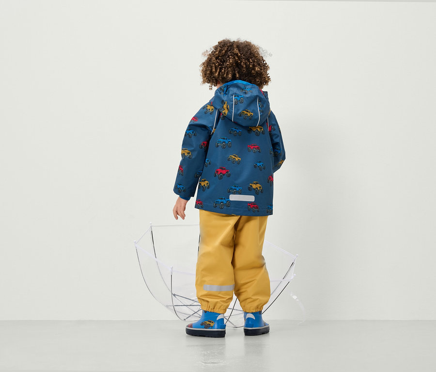 Sırtı kameraya dönük olan bir çocuk, araba baskılı bir ceket ve şeffaf bir şemsiyenin yanında sarı pantolon giyiyor.