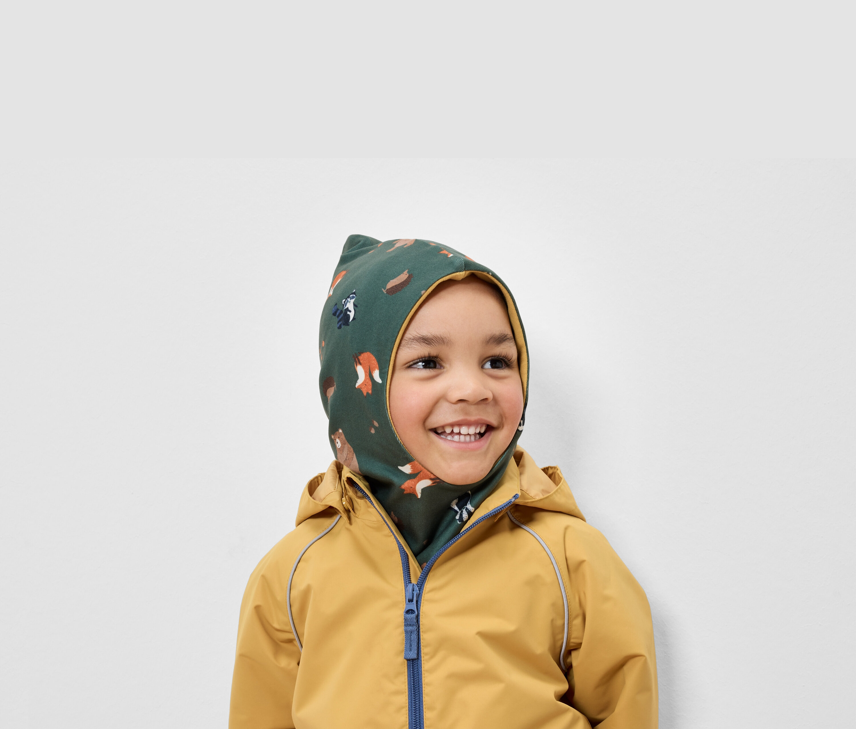 Gülümseyen bir çocuk, hayvan desenli yeşil bir balaklava ve sarı bir ceket giyiyor.