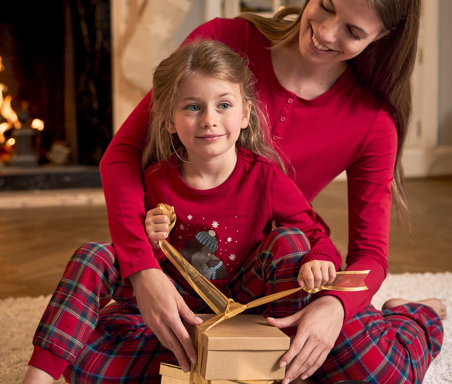 Anne ve kızı halıda oturmuş, altın kurdeleyle Noel hediyeleri paketliyor. İkisi de kırmızı pijama giymiş.