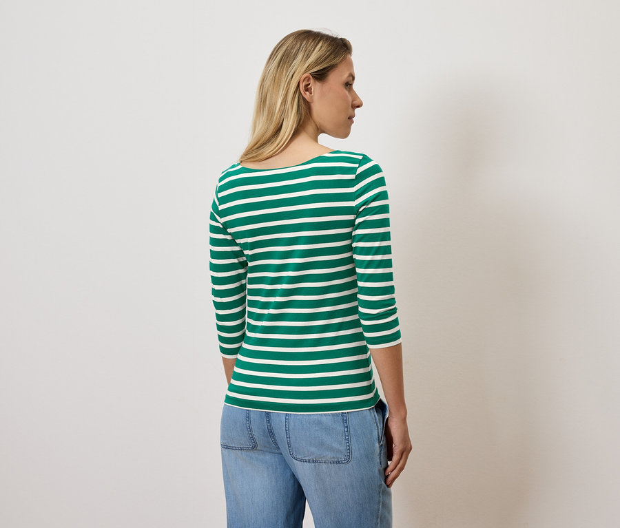Sarışın saçlı bir kadın kameraya arkası dönük duruyor, üzerinde yeşil ve beyaz çizgili üç çeyrek kollu bir tişört ve mavi kot pantolon var.