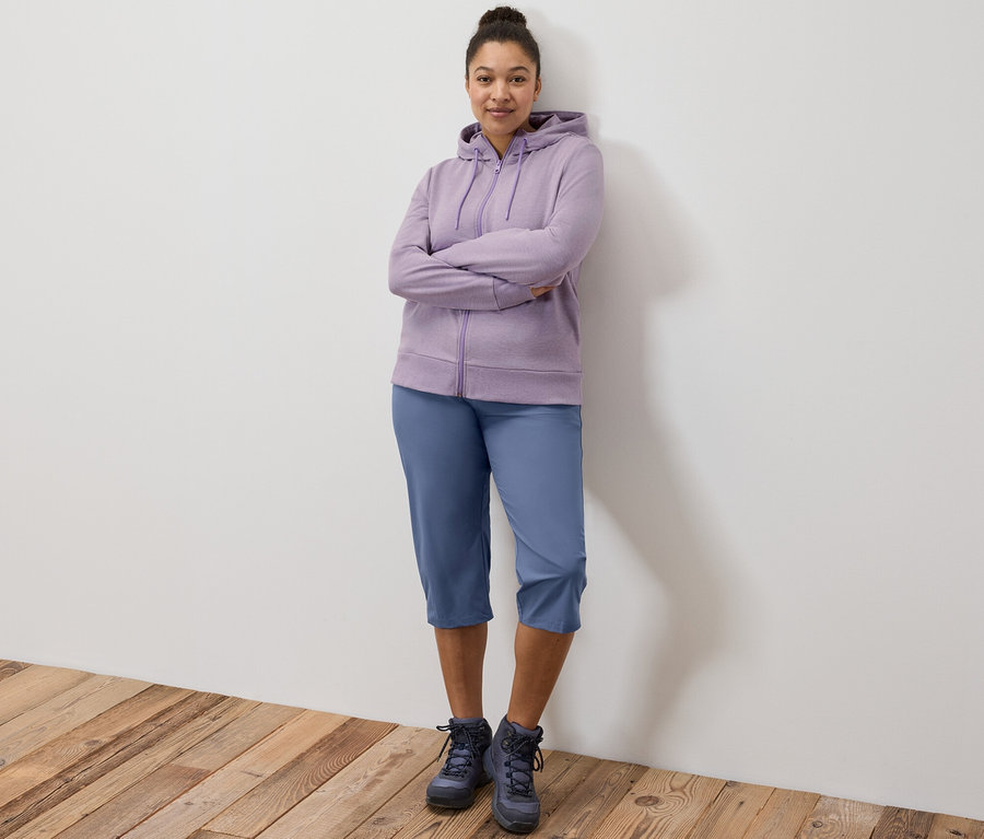 Kollarını kavuşturmuş bir şekilde duran kadın, lila rengi bir sweatshirt ve fonksiyonel kapri giyiyor.