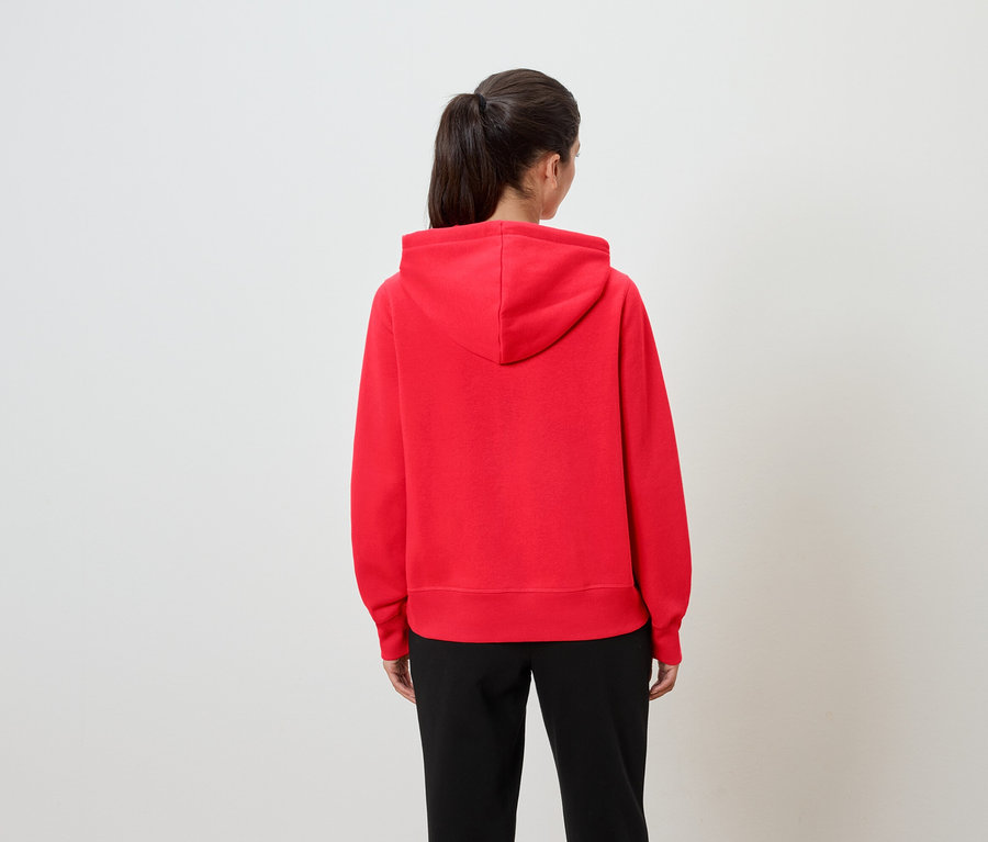 Kadın kameraya arkası dönük, kırmızı kapüşonlu bir sweatshirt ve siyah pantolon giyiyor.