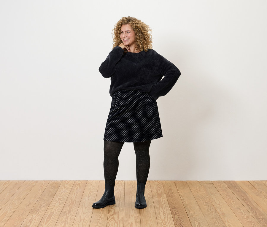 Kıvırcık saçlı bir kadın, siyah bir kazak, puantiyeli etek, külotlu çorap ve botlarla poz veriyor.