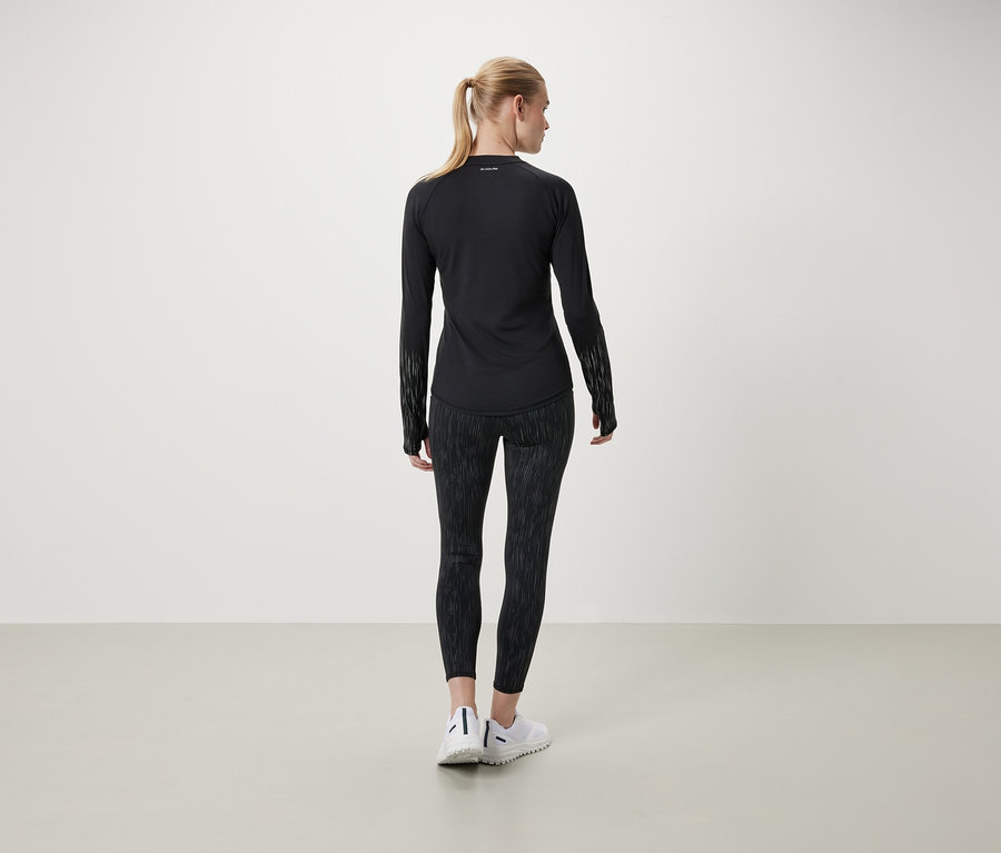 Sırtı kameraya dönük bir kadın, uzun kollu siyah bir tişört ve desenli bir tayt giyiyor.