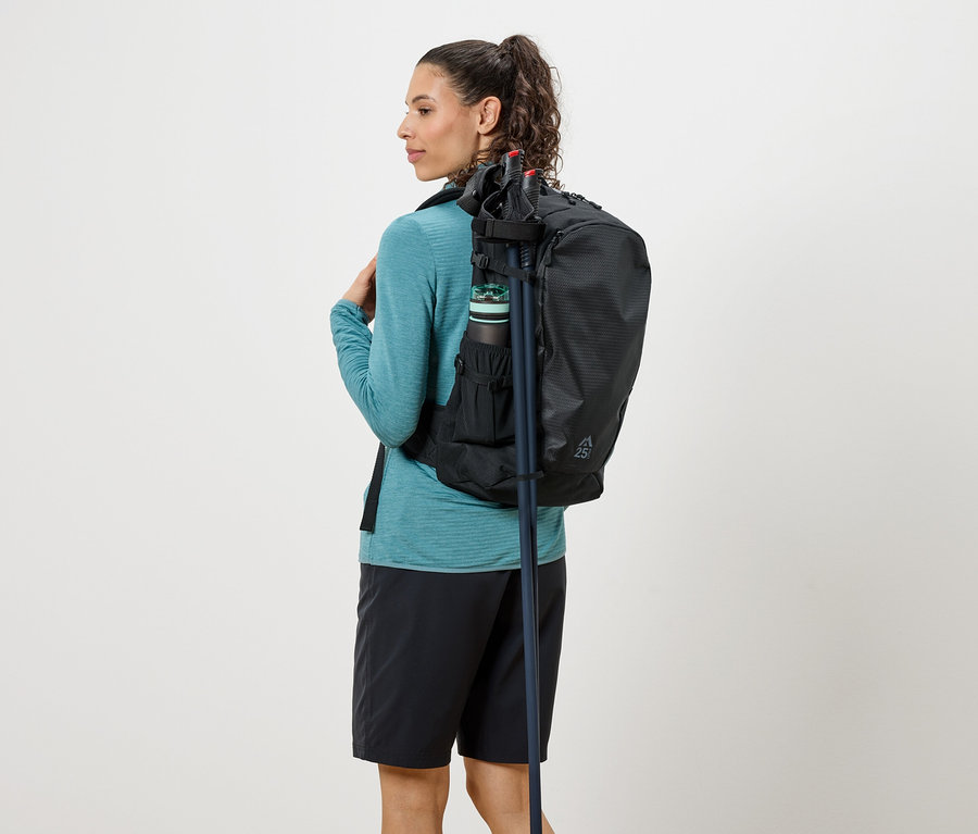 Turkuaz bir üst ve siyah şort giyen, sırtında siyah bir sırt çantası, su şişesi ve yürüyüş sopaları olan bir kadın, arkadan.