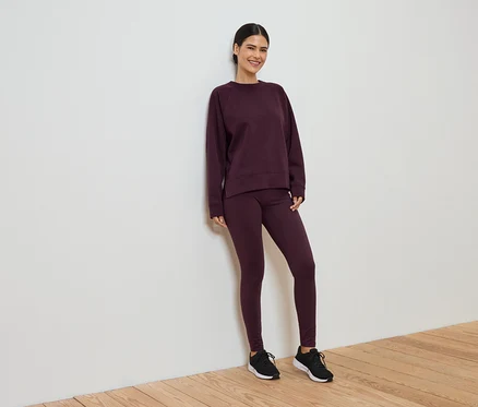 Bordo tayt ve sweatshirt'ten oluşan eşofmanıyla gülümseyen kadın, beyaz duvara yaslanıyor.