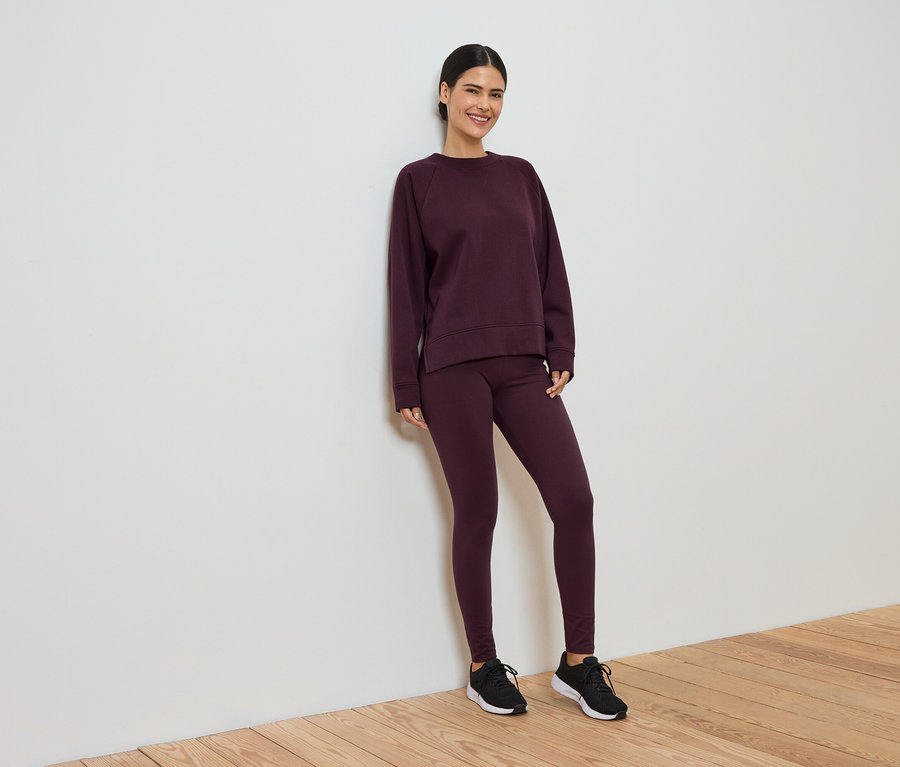 Bordo tayt ve sweatshirt'ten oluşan eşofmanıyla gülümseyen kadın, beyaz duvara yaslanıyor.