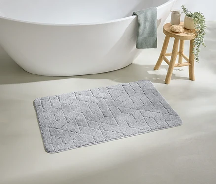 Beyaz bir küvetin önünde geometrik desenli gri bir banyo paspası duruyor. Küvetin kenarından yeşil bir havlu sarkıyor.
