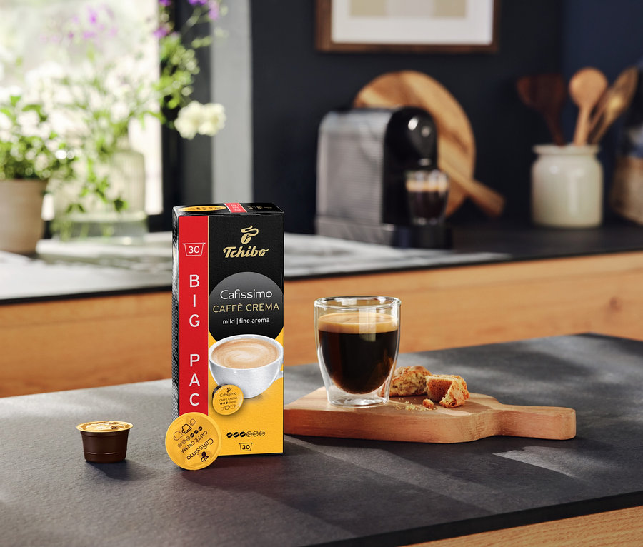 Mutfak tezgahında bir paket Cafissimo Caffè Crema mild kapsül, bir fincan kahve ve bir kahve makinesi.