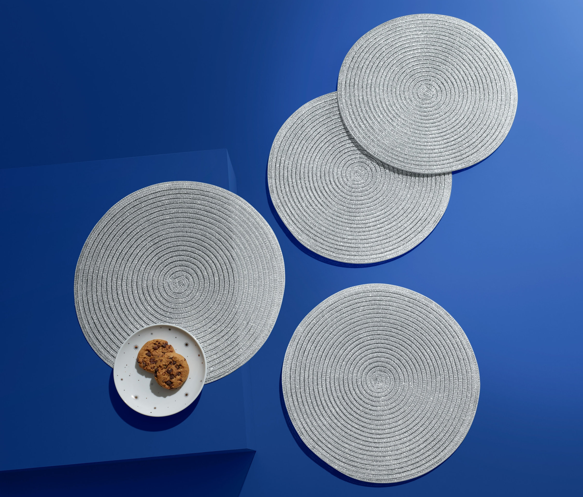 Mavi bir zemin üzerinde dört adet gümüş rengi, yuvarlak Amerikan servis; birinin üzerinde kurabiyelerle dolu bir tabak bulunuyor.