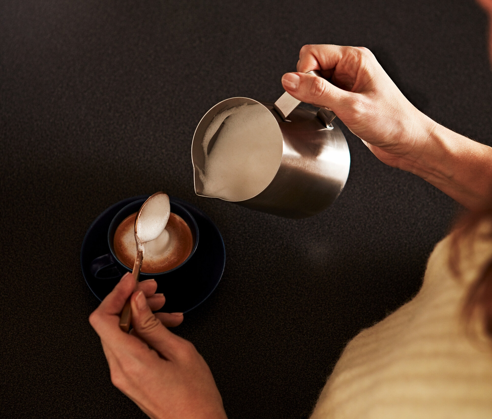 Kadın, küçük paslanmaz çelik sütlükten bir fincan kahveye köpüklü süt döküyor ve kaşıkla köpük alıyor.