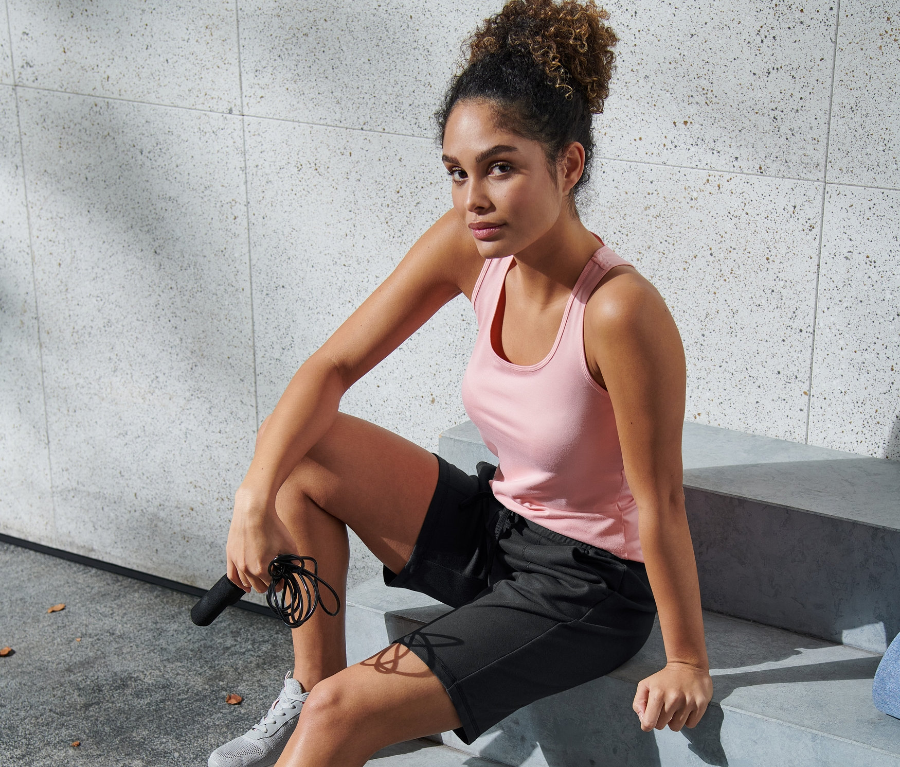 Kadın kahverengi siyah fonksiyonel şortla merdivende oturuyor ve atlama ipi tutuyor.