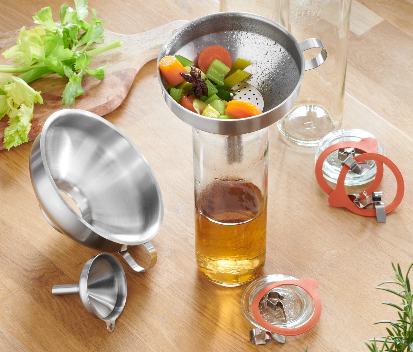Paslanmaz çelik huni seti, kereviz, havuç, bardak ve baharatlarla birlikte ahşap bir masa üzerinde yer almaktadır.