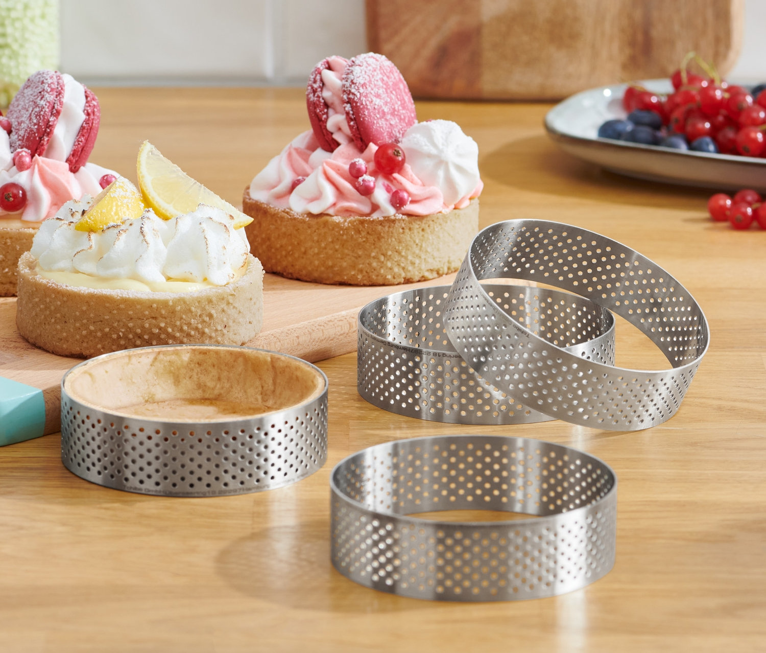 Ahşap bir masa üzerinde, bir tabak meyve ve üç tartın yanında dört adet paslanmaz çelik yuvarlak kalıp.