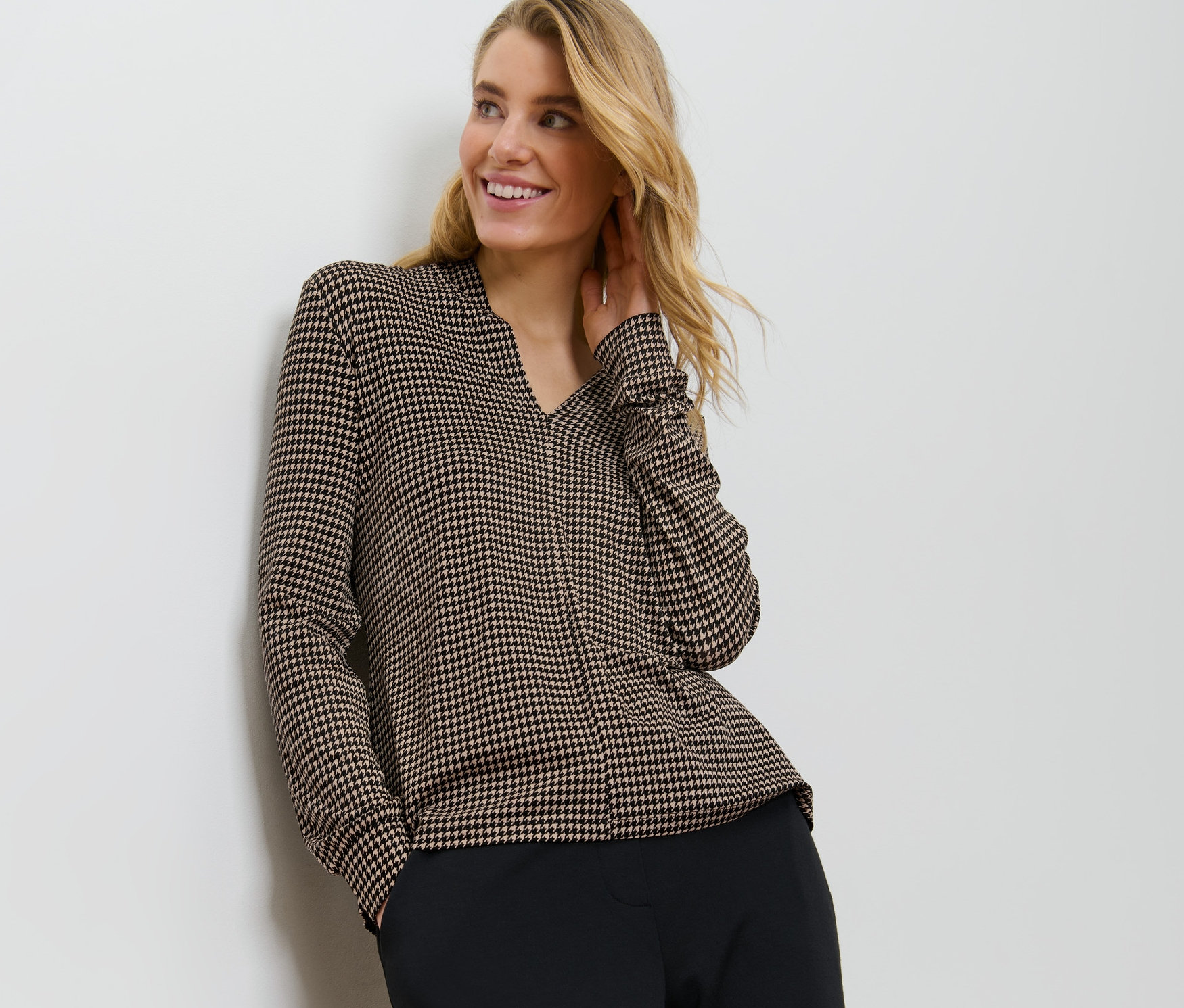 Duvara yaslanmış sarışın bir kadın, V yakalı baskılı bir bluz giyiyor.