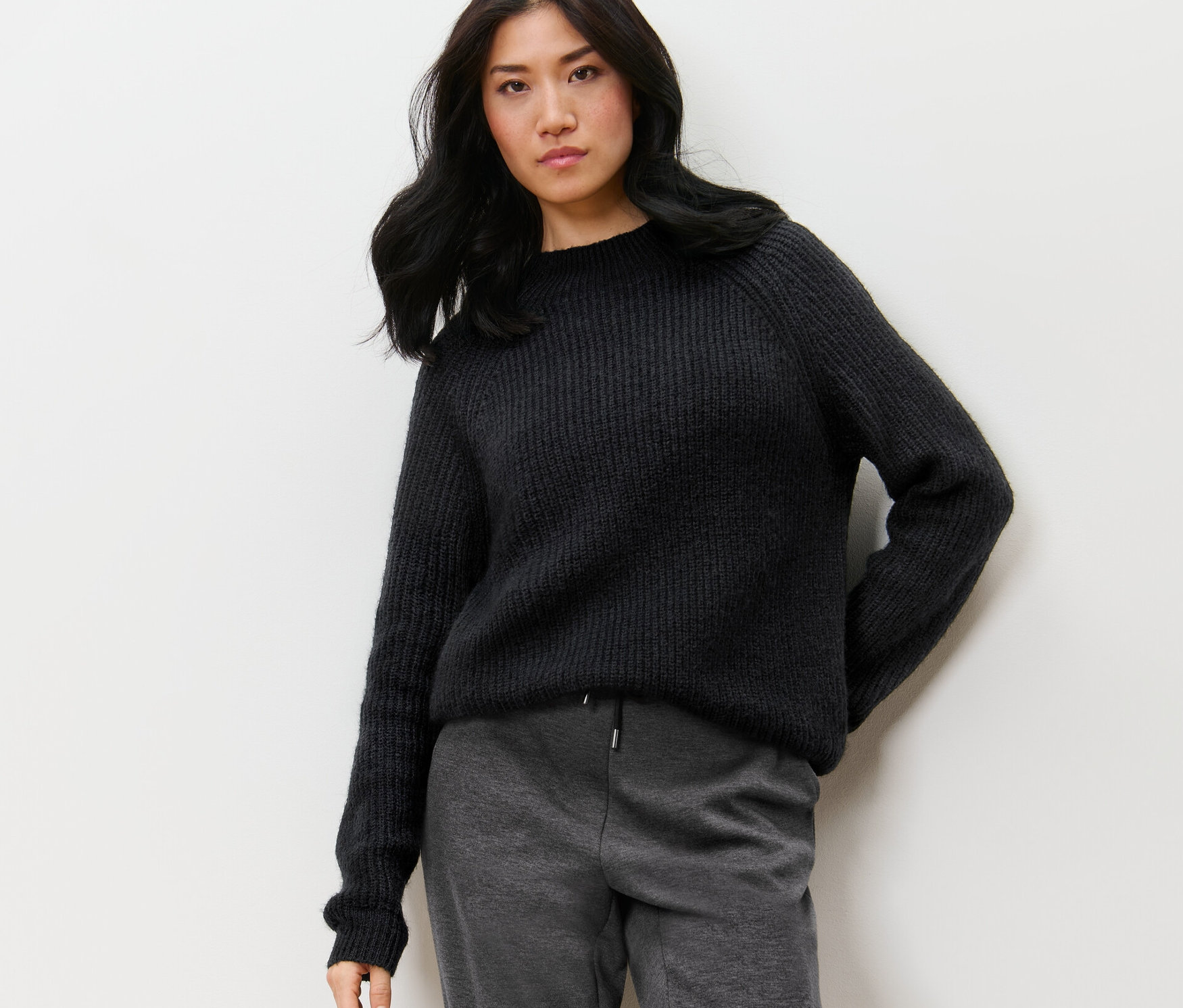 Bir kadın duvar önünde poz veriyor ve siyah, yarım boğazlı kalın örgü bir kazak giyiyor.