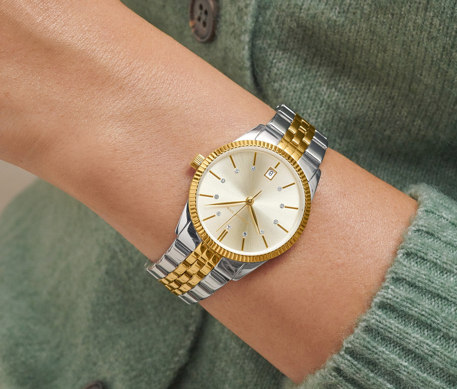 Yeşil bir kazak giyen bir kadının bileğindeki tarihli iki renkli kadın saatinin yakın çekimi.