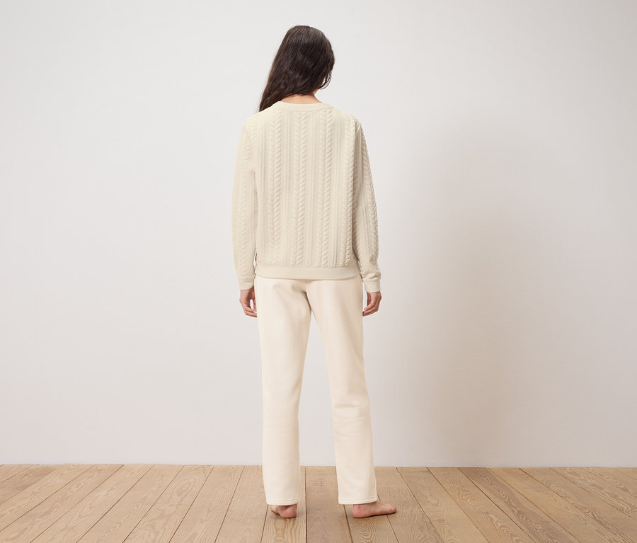 Bir kadın kameraya arkası dönük duruyor, üzerinde krem rengi bir kazak ve pantolon var.