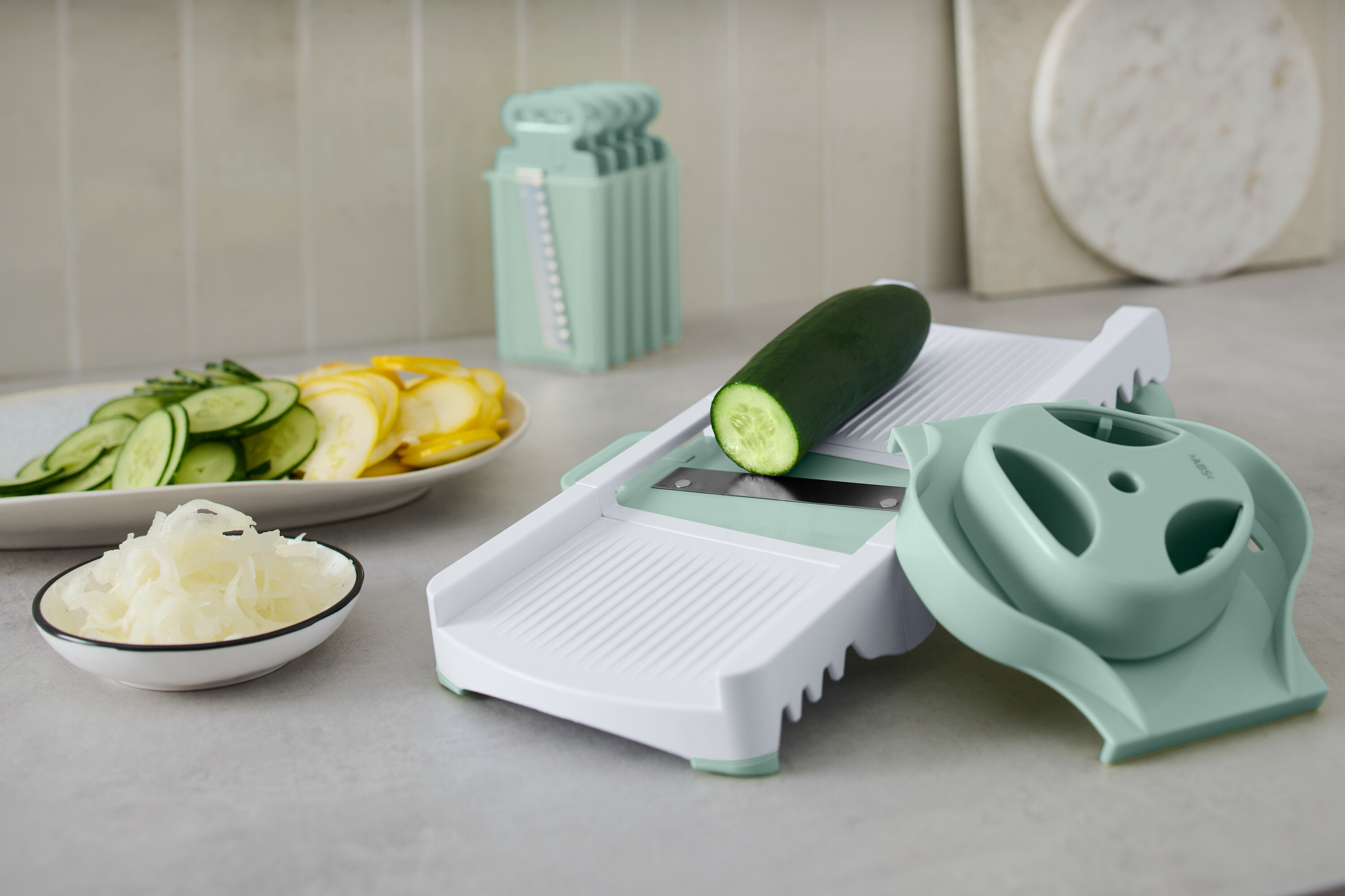 Beyaz bir sebze dilimleyicide aksesuarlarıyla birlikte duran yeşil salatalık. Tabakta dilimlenmiş salatalık ve sarı kabak var.