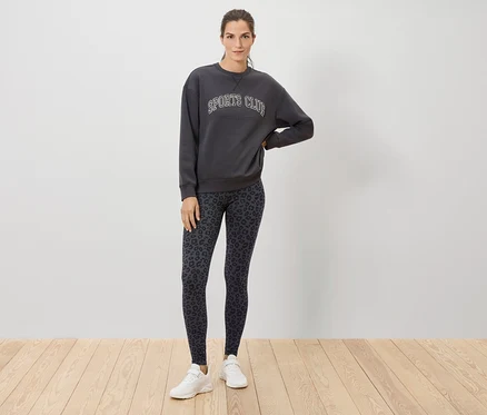 Bir kadın tahta zeminde duruyor, üzerinde SPORTS CLUB yazılı gri bir sweatshirt, hayvan desenli tayt ve beyaz spor ayakkabılar var.