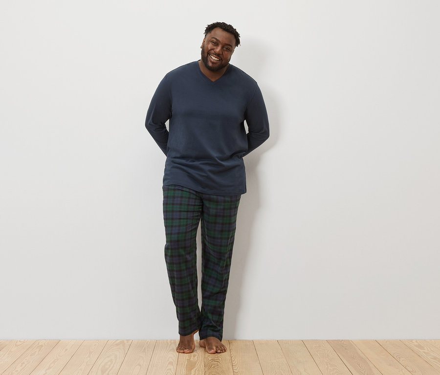 Esmer tenli bir adam ayakta duruyor ve kameraya gülümsüyor. Üzerinde uzun kollu lacivert bir pijama üstü ve ekose pijama altı var.