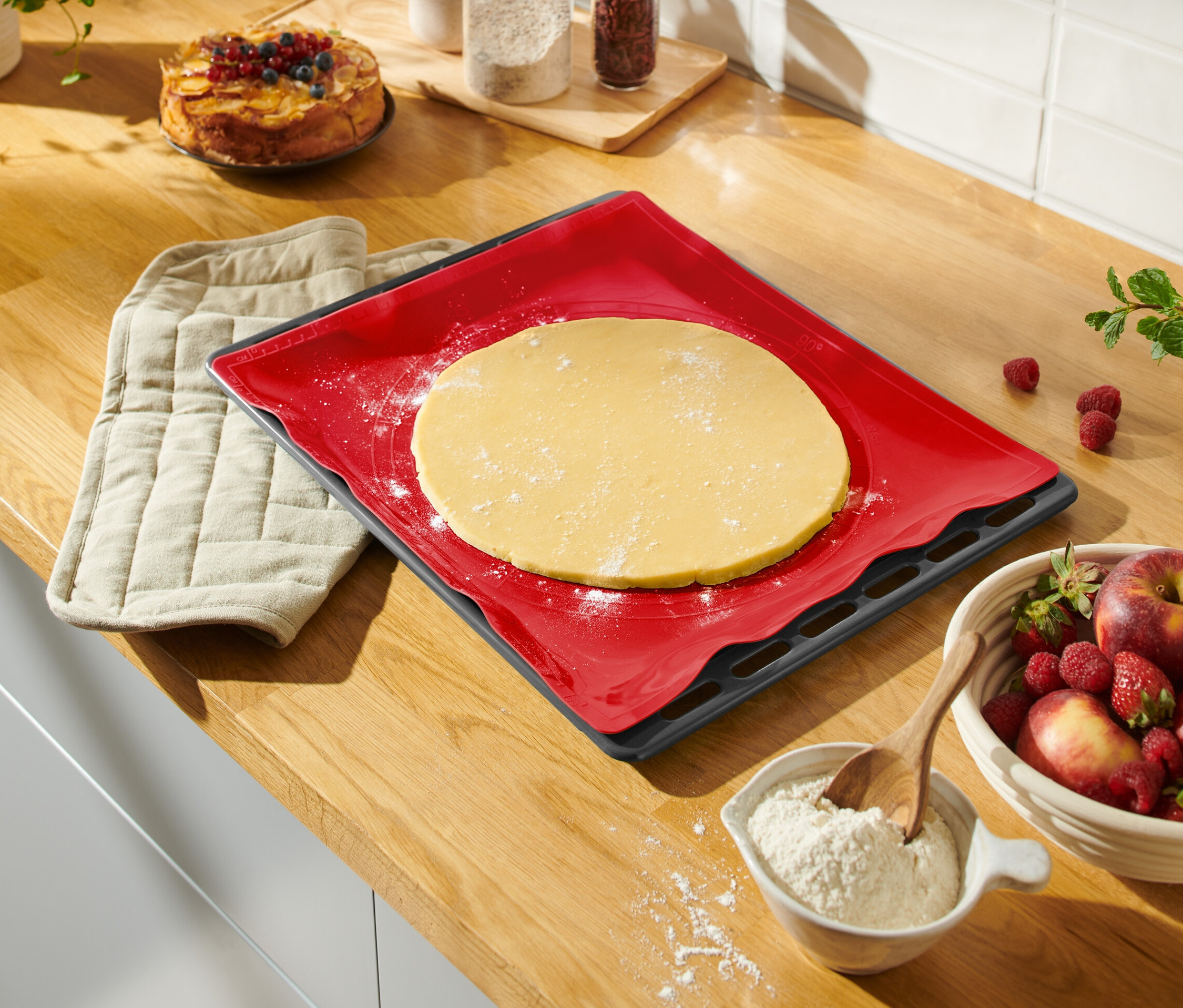 Tezgahta, açılmış hamurlu kırmızı bir pişirme matı, bir tutacak, bir kase meyve ve bir kek var.