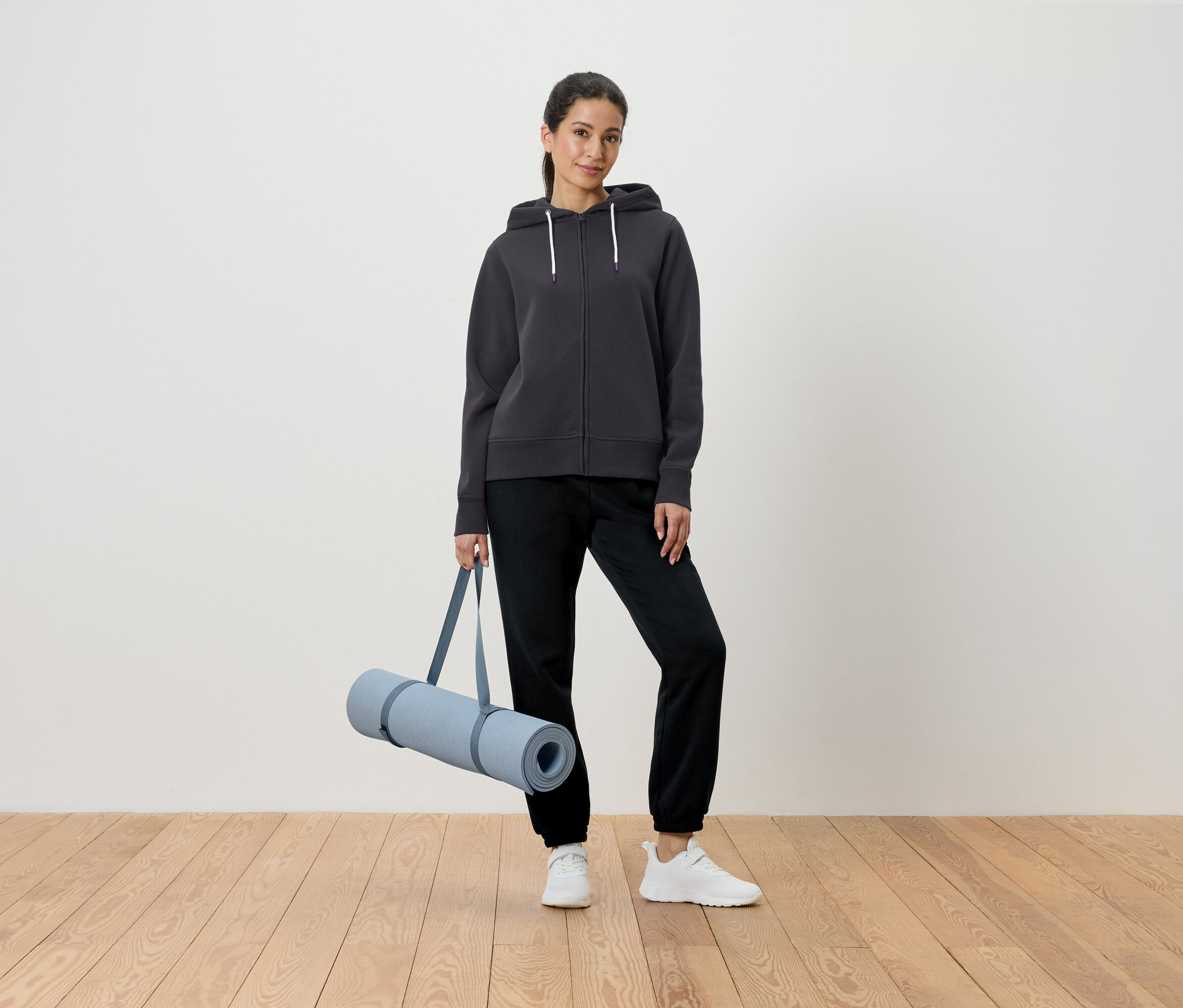 Kadın gri kapüşonlu ve siyah pantolonla yoga matı tutuyor.