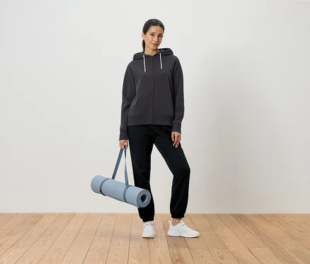 Kadın gri kapüşonlu ve siyah pantolonla yoga matı tutuyor.