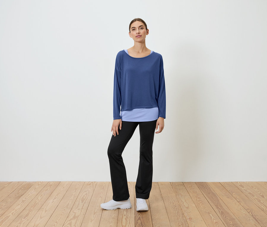 Bir kadın ahşap bir zeminde duruyor, üzerinde mavi bir bluz ve siyah bir pantolon var.