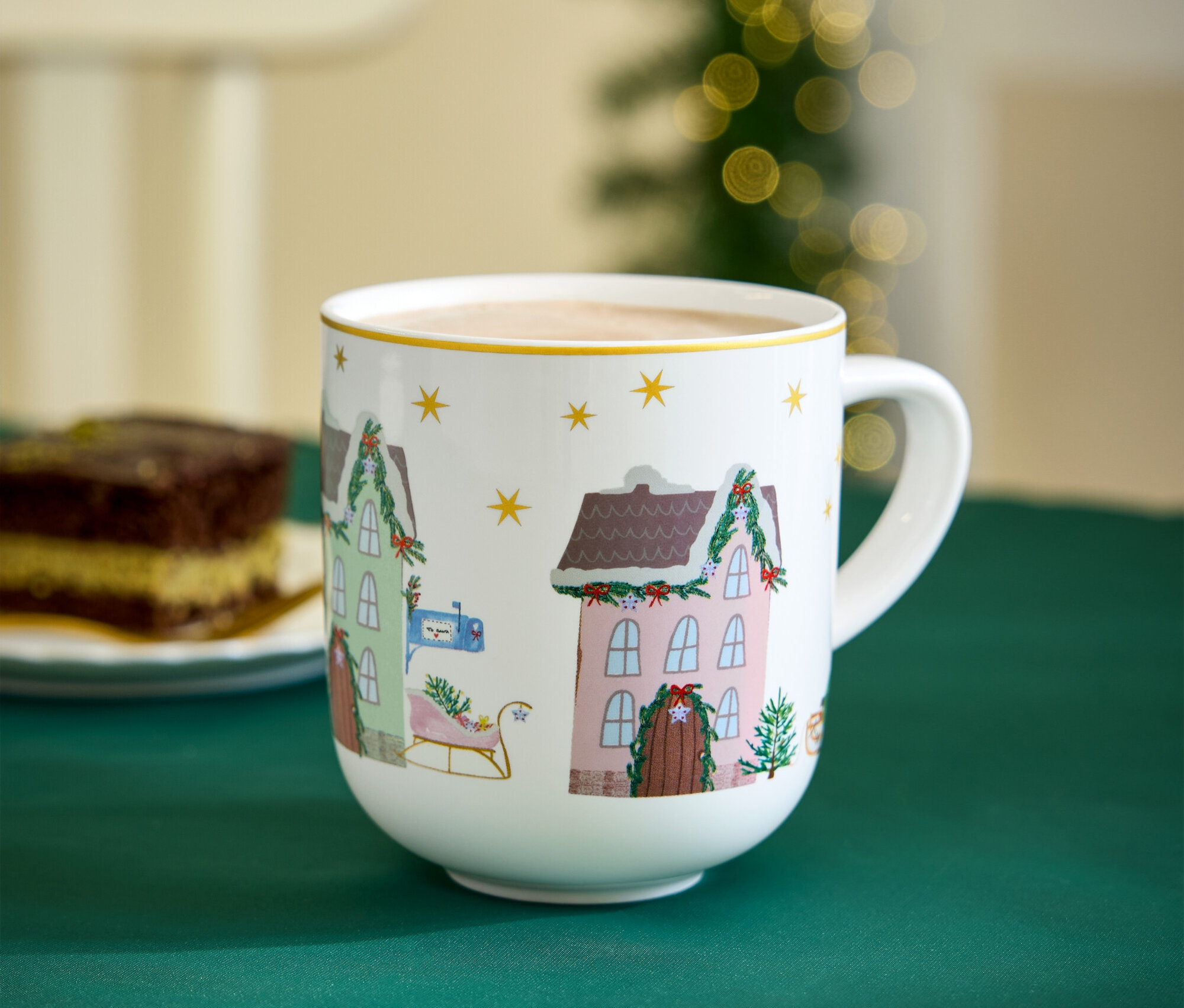 Noel temalı bir kupa, yeşil bir örtü üzerinde bir dilim kekin yanında duruyor.