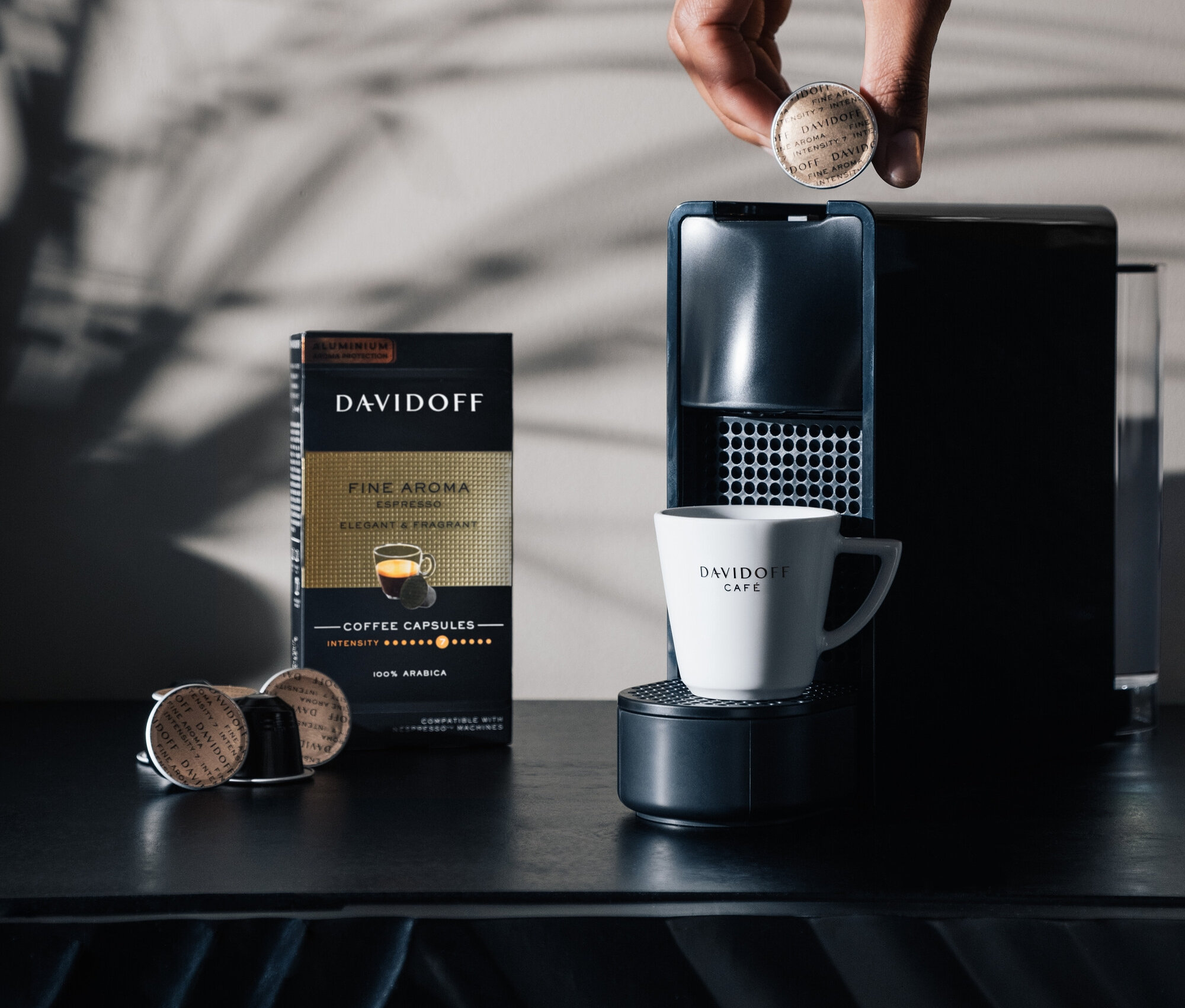 Bir el Davidoff kahve makinesine bir kapsül yerleştiriyor. Yanında bir paket Davidoff Fine Aroma kapsülü ve bir Davidoff Cafe fincanı duruyor.