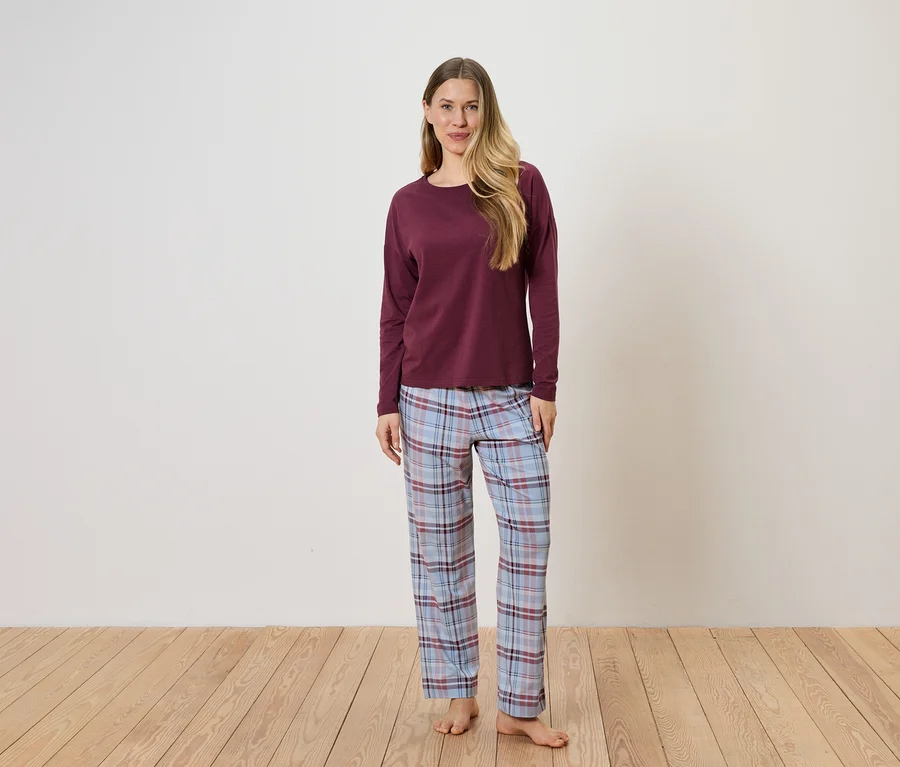Kadın beyaz duvar önünde duruyor, bordo uzun kollu bir bluz ve mavi ekose pijama altı giyiyor.
