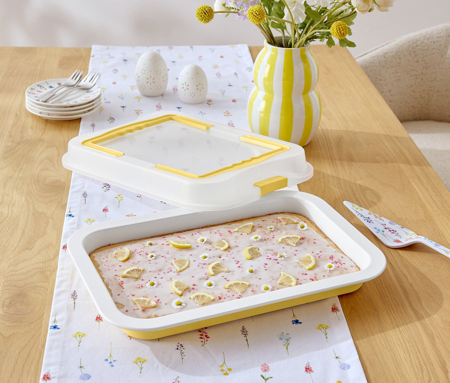 Ahşap bir masanın üzerinde limonlu sır ve papatyalarla süslenmiş bir kek, sarı ve beyaz çizgili bir sürahi, çiçek desenli bir pasta spatulası ve çiçek desenli beyaz bir masa örtüsü bulunmaktadır.