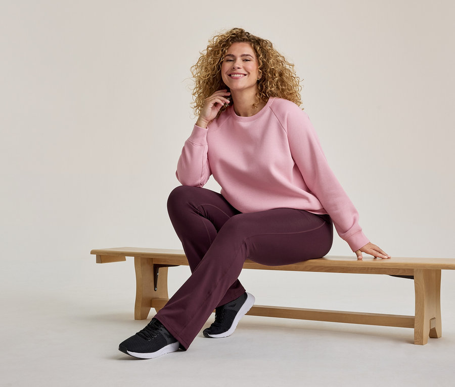 Kıvırcık saçlı, pembe sweatshirt ve koyu mor pantolon giymiş, ahşap bir bankta oturan gülümseyen kadın.