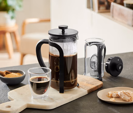 Siyah French Press 800 ml, 6 Fincan, kahve dolu, mermer bir tahta üzerinde bir bardak kahve, bir kase şeker ve kurabiyelerin yanında duruyor. Yanında demonte edilmiş başka bir French Press var.