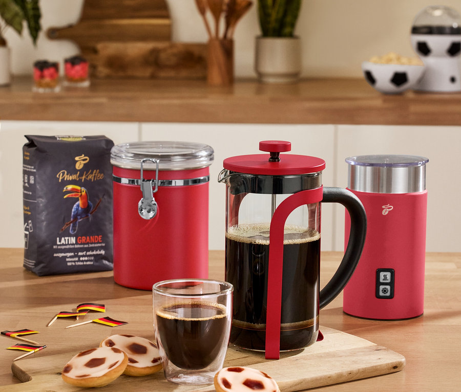 Tezgahta kırmızı aroma kutusu, kırmızı elektrikli süt köpürtücü, kırmızı french press, 2 adet çift cidarlı caffè crema bardağı ve bir paket kahve duruyor.