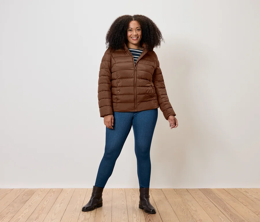 Kıvırcık saçlı, gülümseyen bir kadın, konyak rengi kapitone mont, mavi kot pantolon ve koyu renk botlar giymiş, açık renk duvar ve ahşap zemin önünde duruyor.