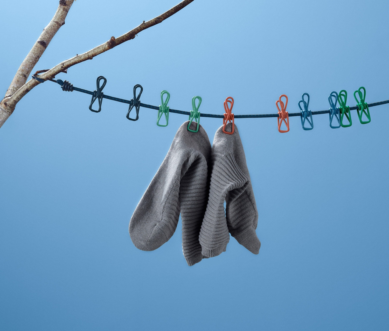 Mavi bir arka planın önünde, bir ağacın dalına tutturulmuş bir kamp çamaşır ipinde asılı duran iki gri çorap.