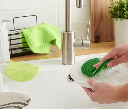 Kadın, beyaz bir lavaboda yeşil 2 Adet Silikon Bulaşık Pedi ile bir tabağı yıkıyor.