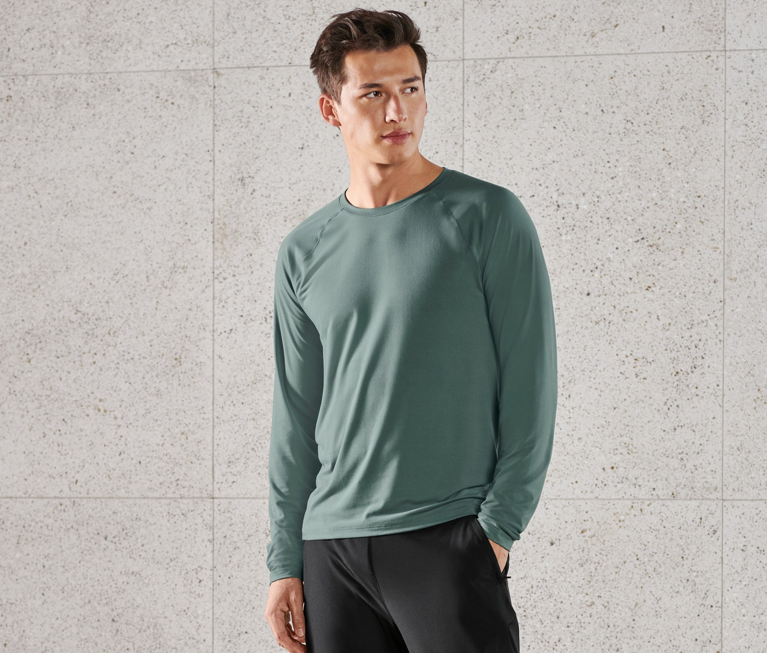 Adam ayakta duruyor ve koyu yeşil uzun kollu fonksiyonel tişört, siyah pantolon ve siyah ayakkabılar giyiyor.