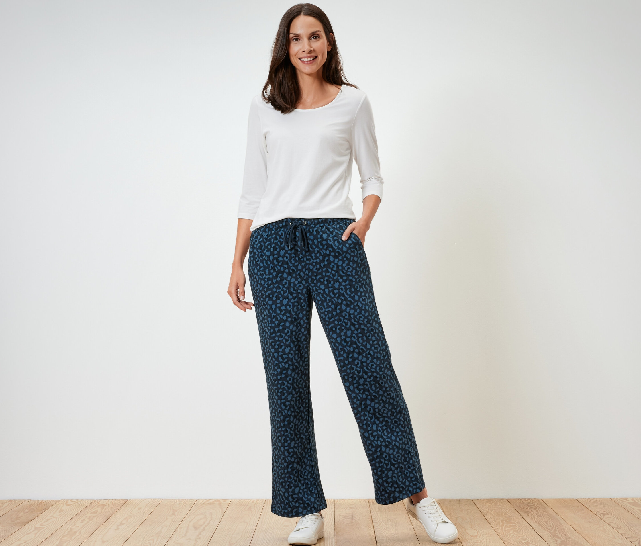 Bir kadın ayakta duruyor, üzerinde beyaz, uzun kollu bir tişört ve baskılı eşofman altı var.