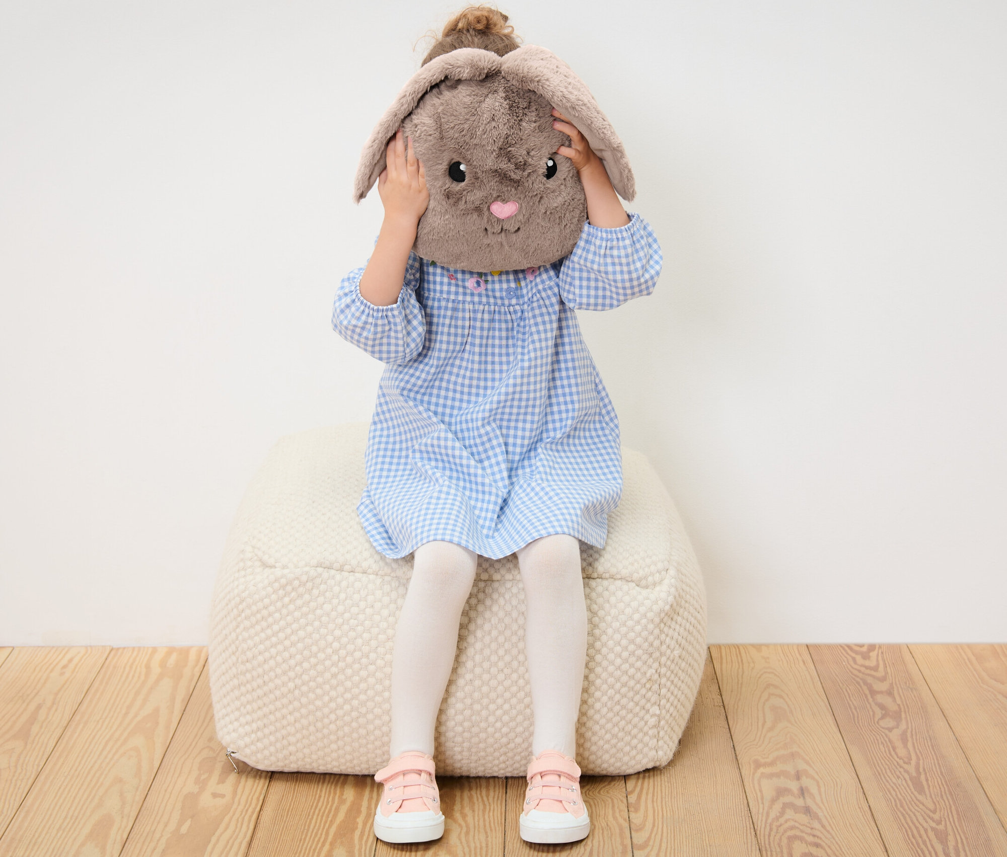 Kız bir pufun üzerinde oturuyor ve yüzünün önünde pelüş tavşan yastık tutuyor.