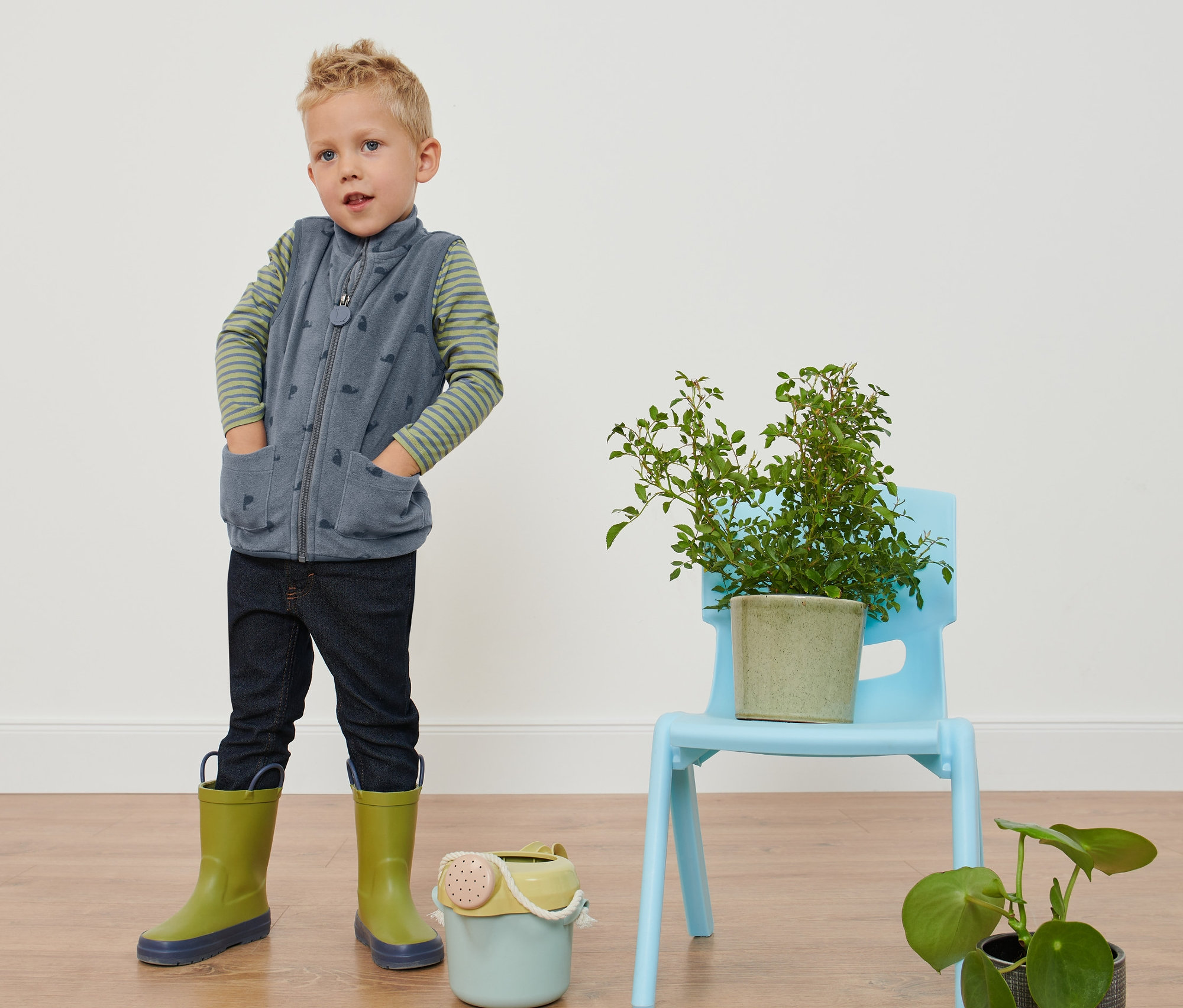 Çocuk elleri cebinde salyangoz baskılı polar yelek, kot pantolon ve lastik çizme ile duruyor.
