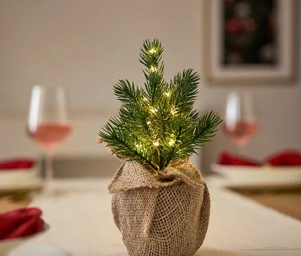 Masa üzerinde örtü, tabak ve bardaklarla birlikte çuval bezi ambalajında ışıklı küçük bir Noel ağacı.